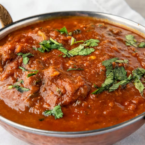 Close-up of a copper bowl filled with rich, red Indian curry, garnished with fresh cilantro.